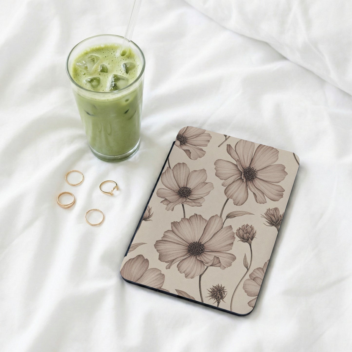Journal with floral pattern, iced matcha latte, and gold rings on white fabric.