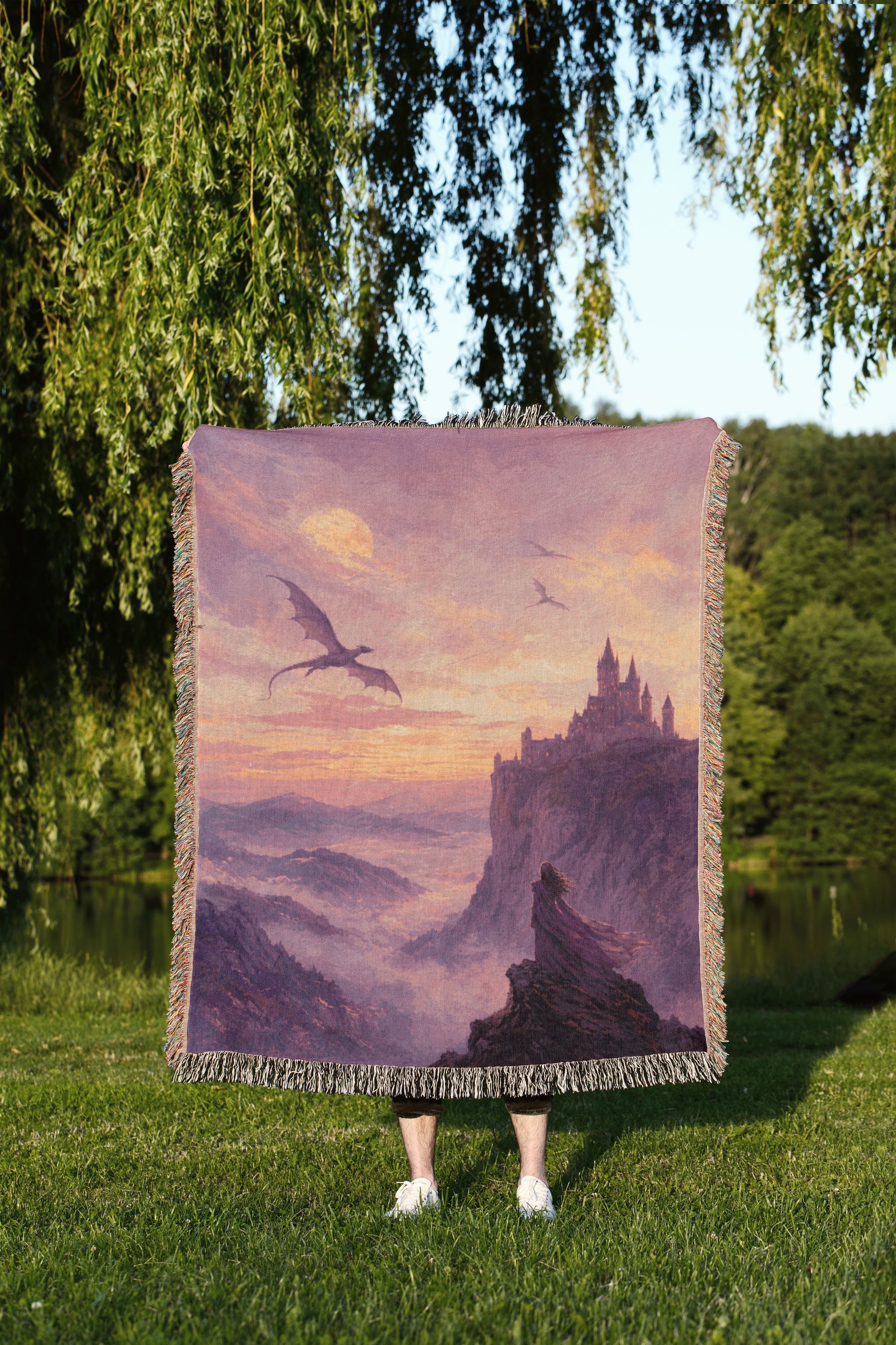 A person is standing in a grassy area, holding a large square blanket that features a painting of a castle and a dragon flying over a mountainous landscape.