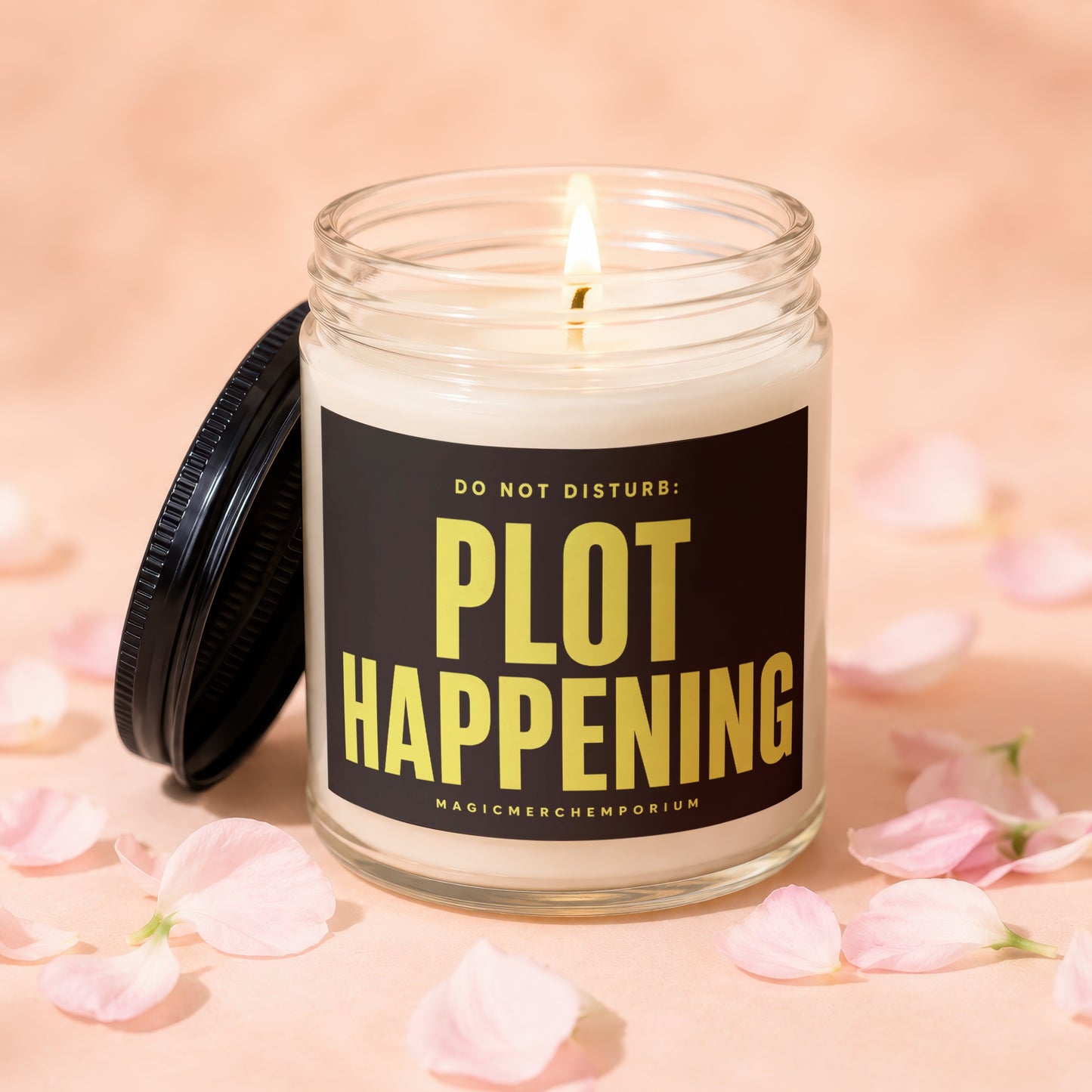 Scented candle with "PLOT HAPPENING" text, lit flame, and soft pink petals.