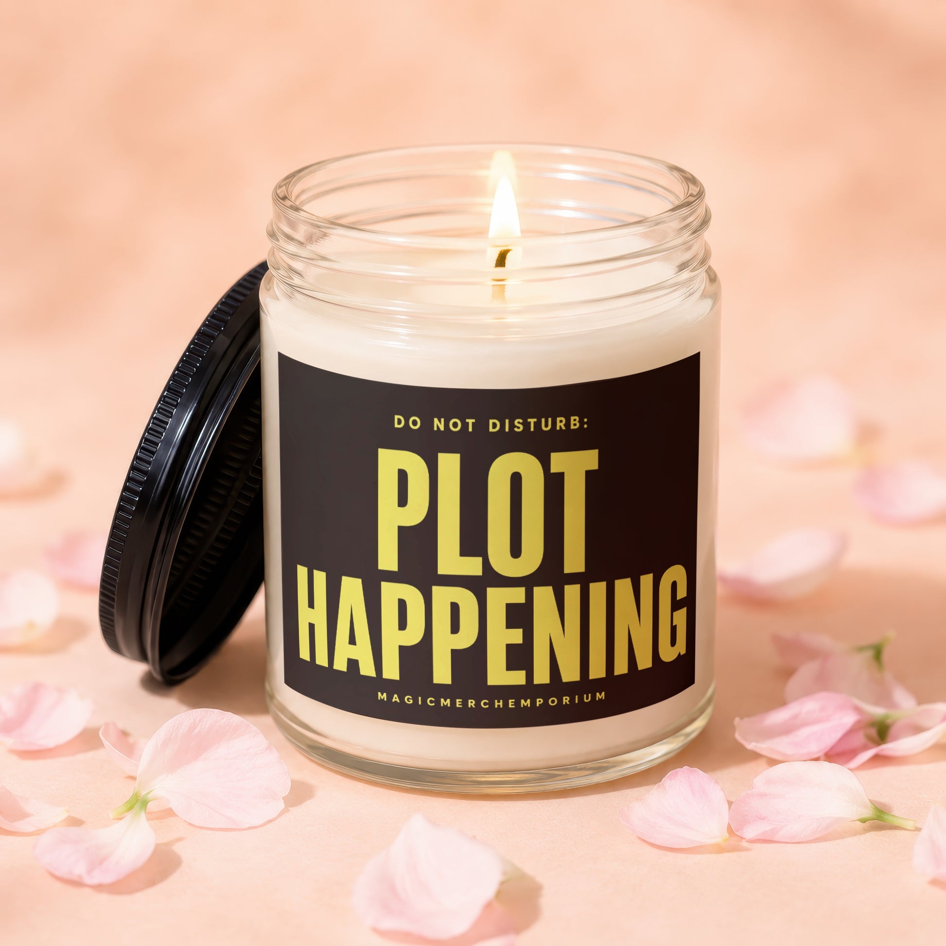 Scented candle with "PLOT HAPPENING" text, lit flame, and soft pink petals.