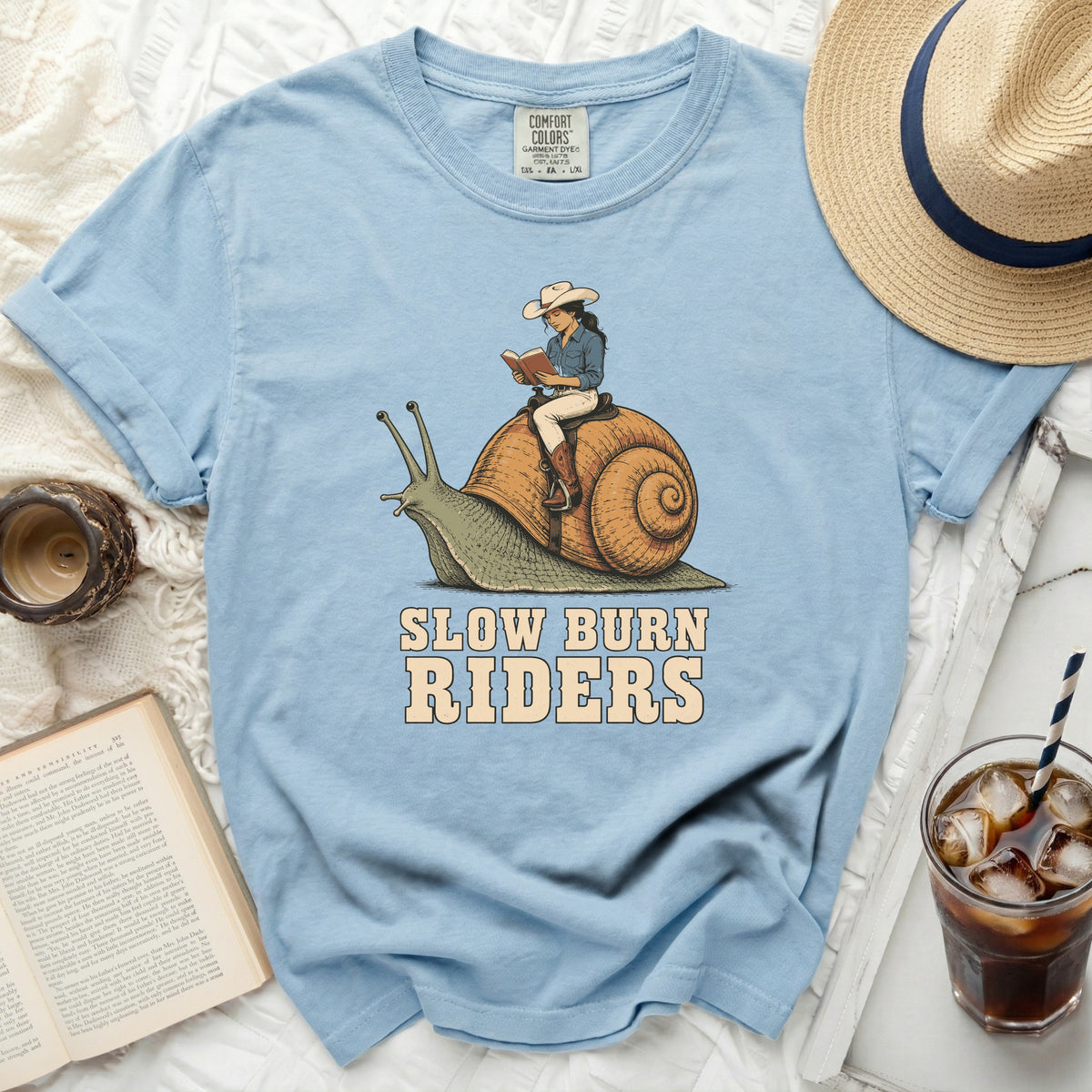 Light blue t-shirt with graphic of woman riding a snail, reading. Text says "SLOW BURN RIDERS."