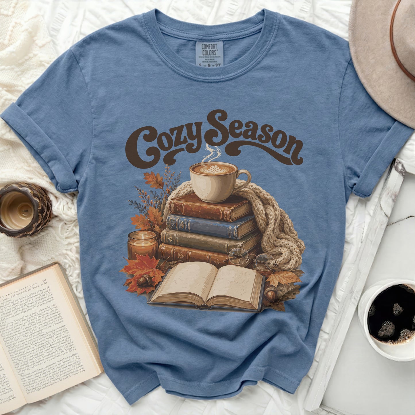 Blue t-shirt with "Cozy Season" text, stacked books, coffee, and autumn leaves.