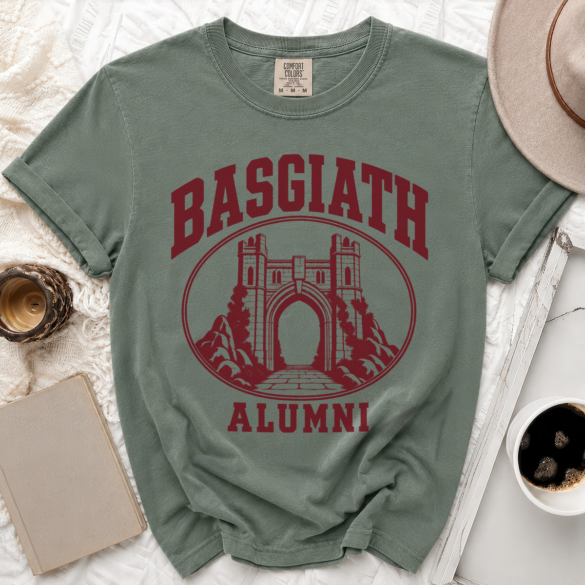 A sage green t-shirt with maroon lettering reading Basgiath Alumni above a castle gate graphic.