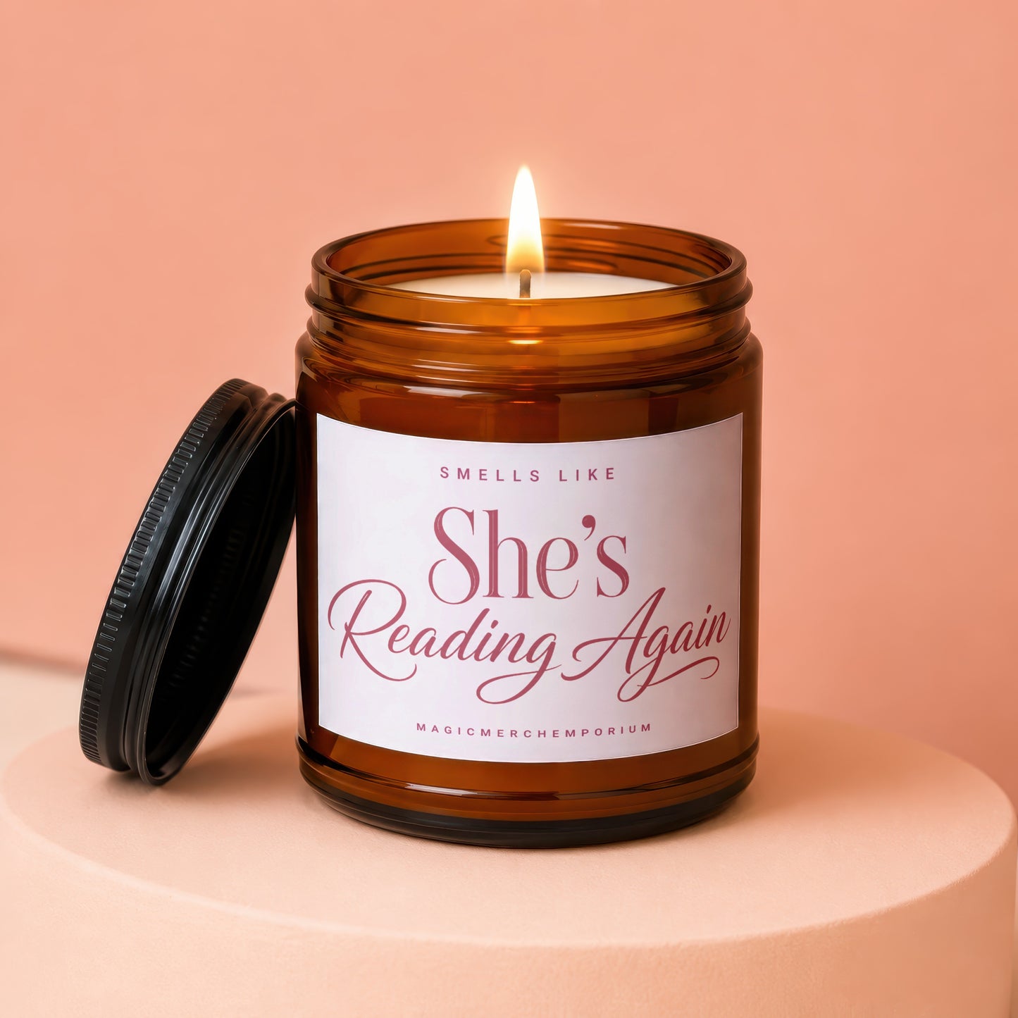 Amber jar candle with lit wick, lid off, labeled "She's Reading Again".