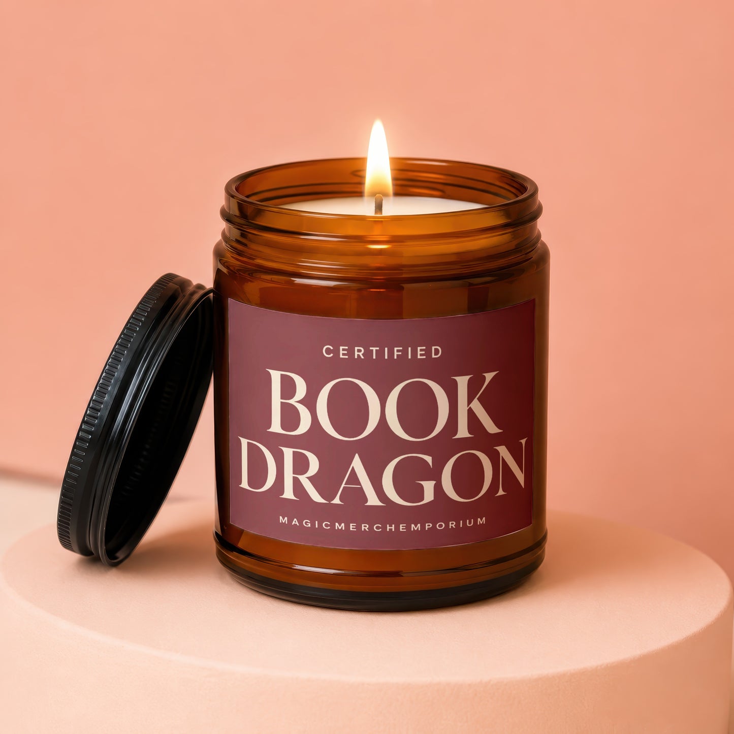 Amber jar candle with lit wick and lid next to it, label reads "Certified Book Dragon".