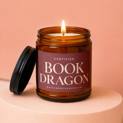 Amber jar candle with lit wick and lid next to it, label reads "Certified Book Dragon".