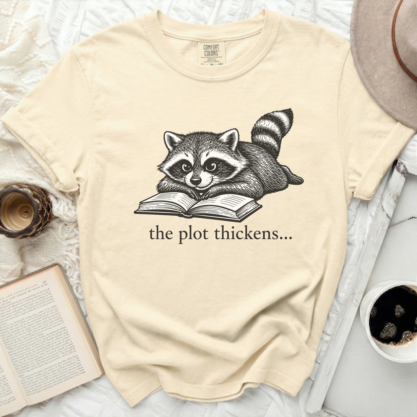 Cream t-shirt with black ink drawing of a raccoon reading a book with text "the plot thickens...".