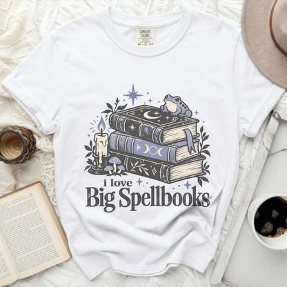 White t-shirt with a graphic of spellbooks, candle, mushrooms, stars, and a frog.