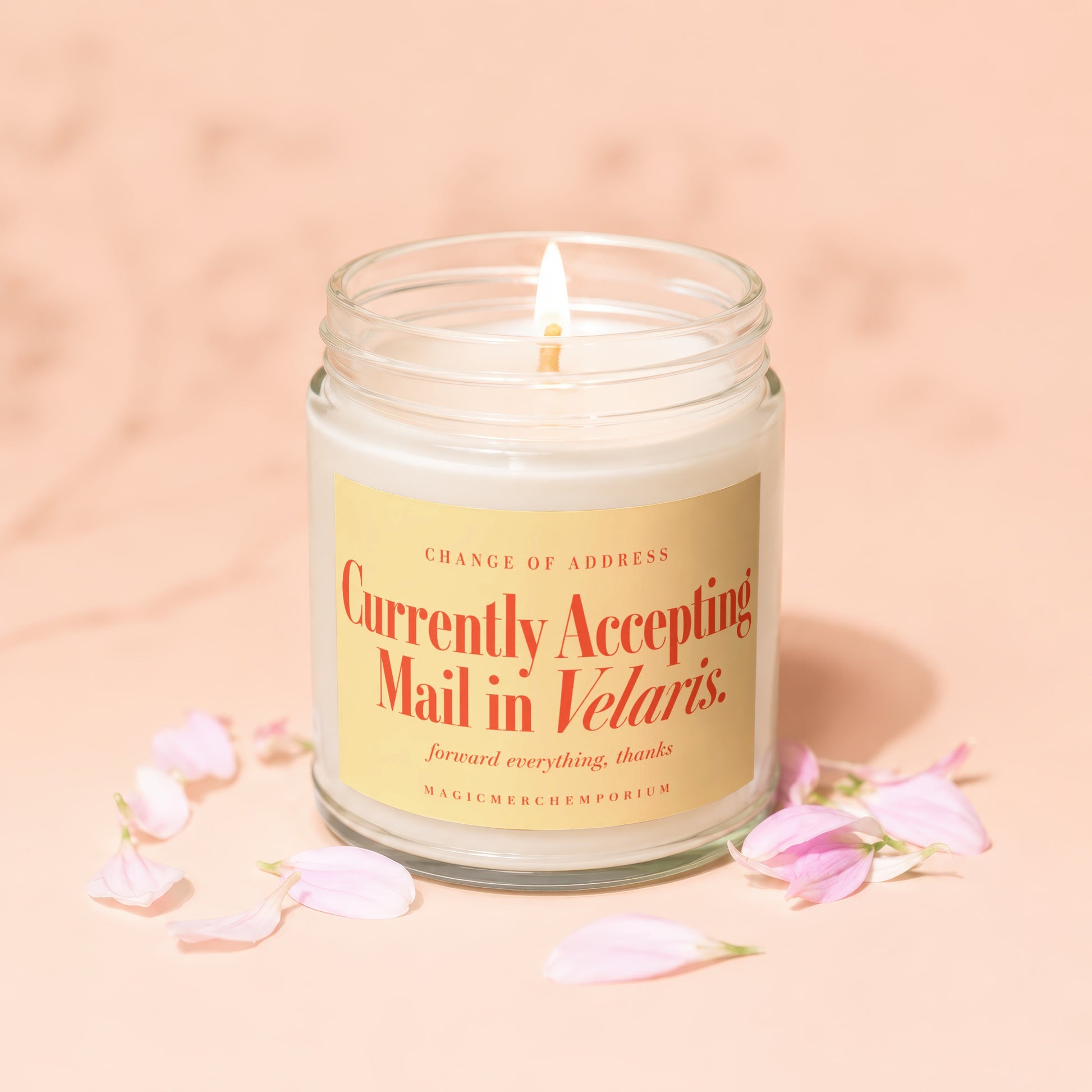 A lit white candle in a glass jar with a yellow label that reads Currently Accepting Mail in Velaris, surrounded by pink petals on a peach background.