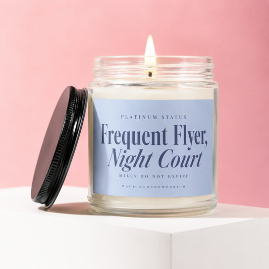 Lit candle in glass jar with "Frequent Flyer, Night Court" label on pink background.