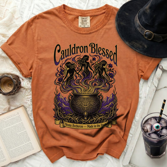 Orange t-shirt with "Cauldron Blessed" graphic, three witches, cauldron, and text.