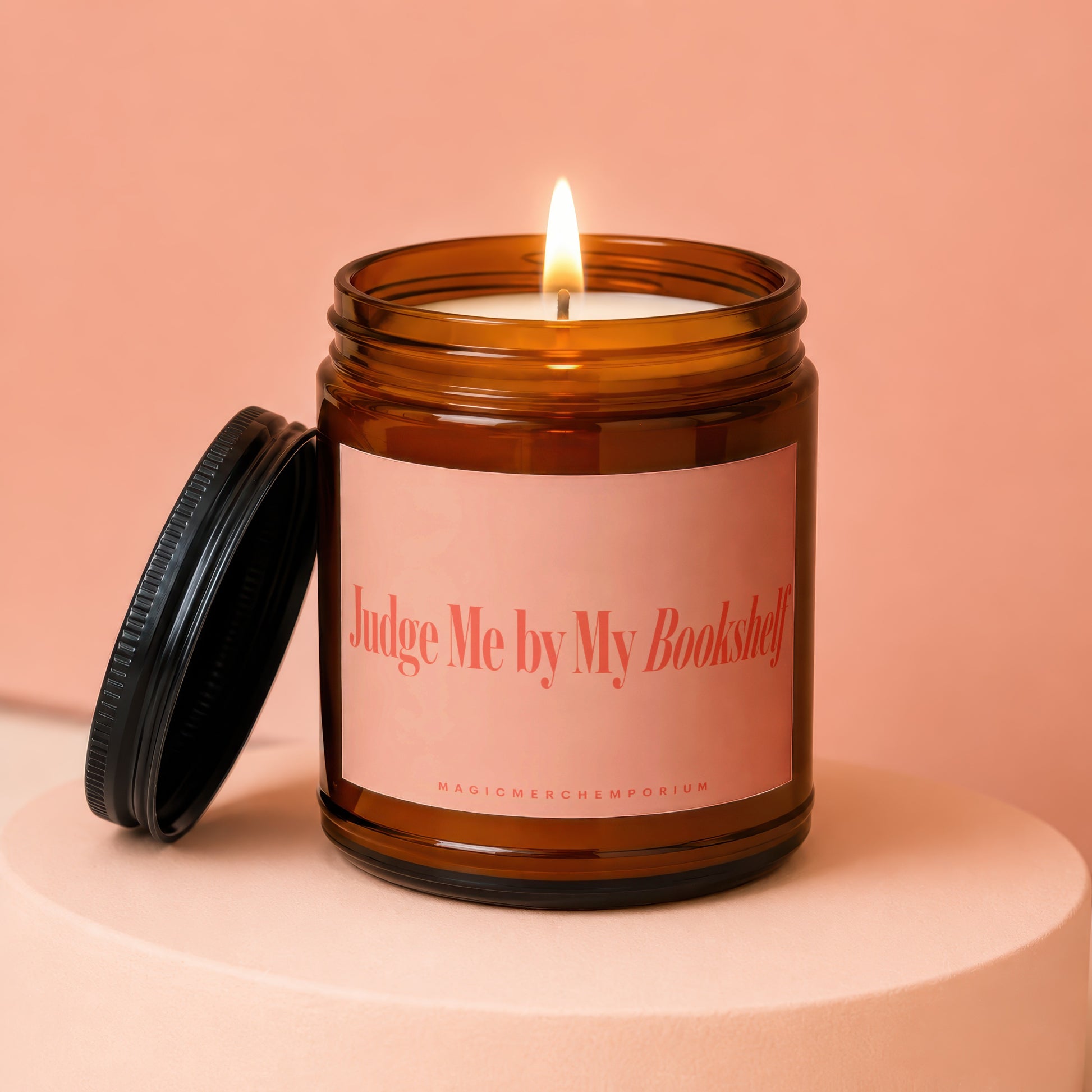 Amber jar candle with lit wick and pink label reading "Judge Me by My Bookshelf"