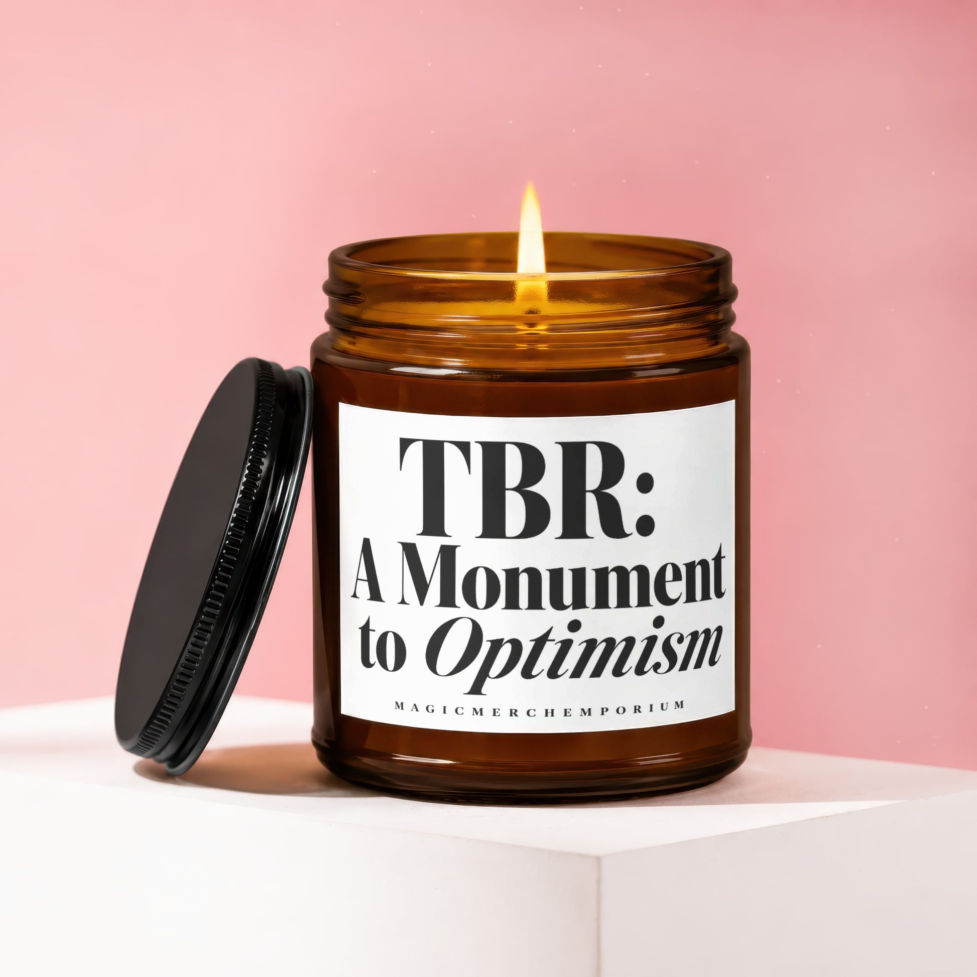 Amber jar candle with lit flame, black lid, and label reading "TBR: A Monument to Optimism".