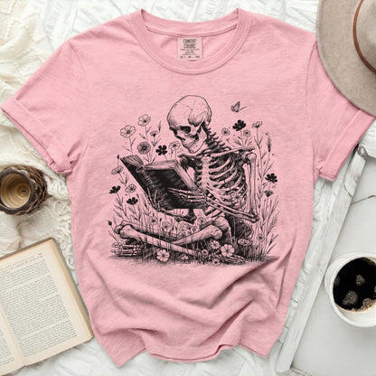 Pink t-shirt with a skeleton reading a book among flowers.