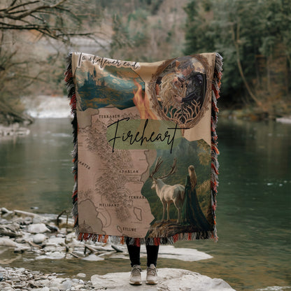 A person is standing on a rock, holding a large blanket with a map and various images of animals, including a deer and a castle, against a backdrop of a river and trees.