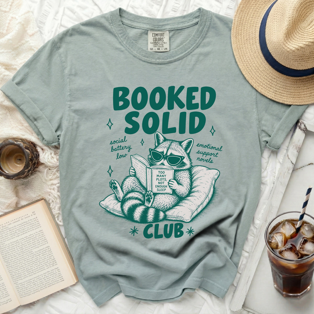 Green t-shirt with "Booked Solid" and a raccoon reading with text "Too Many Plots, Not Enough Sleep Club".