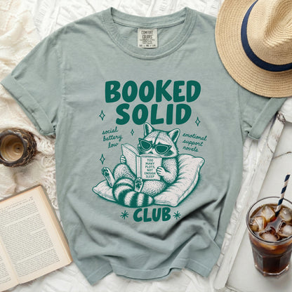 Green t-shirt with "Booked Solid" and a raccoon reading with text "Too Many Plots, Not Enough Sleep Club".