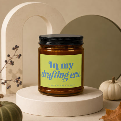 Amber glass candle with black lid and green label reading "In my drafting era."