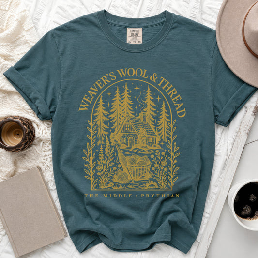 A teal t-shirt featuring a gold graphic of a cabin, pine trees, and text reading "Weaver's Wool & Thread".