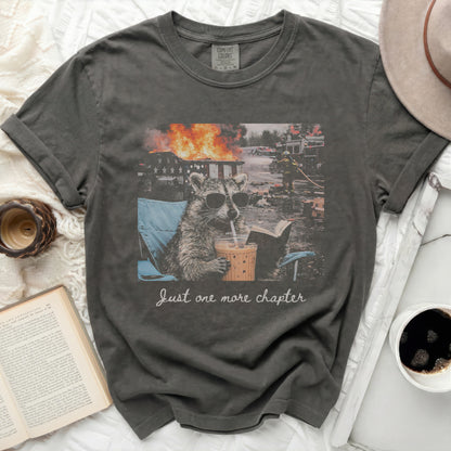 T-shirt with raccoon in sunglasses reading book near fire, text "Just one more chapter".