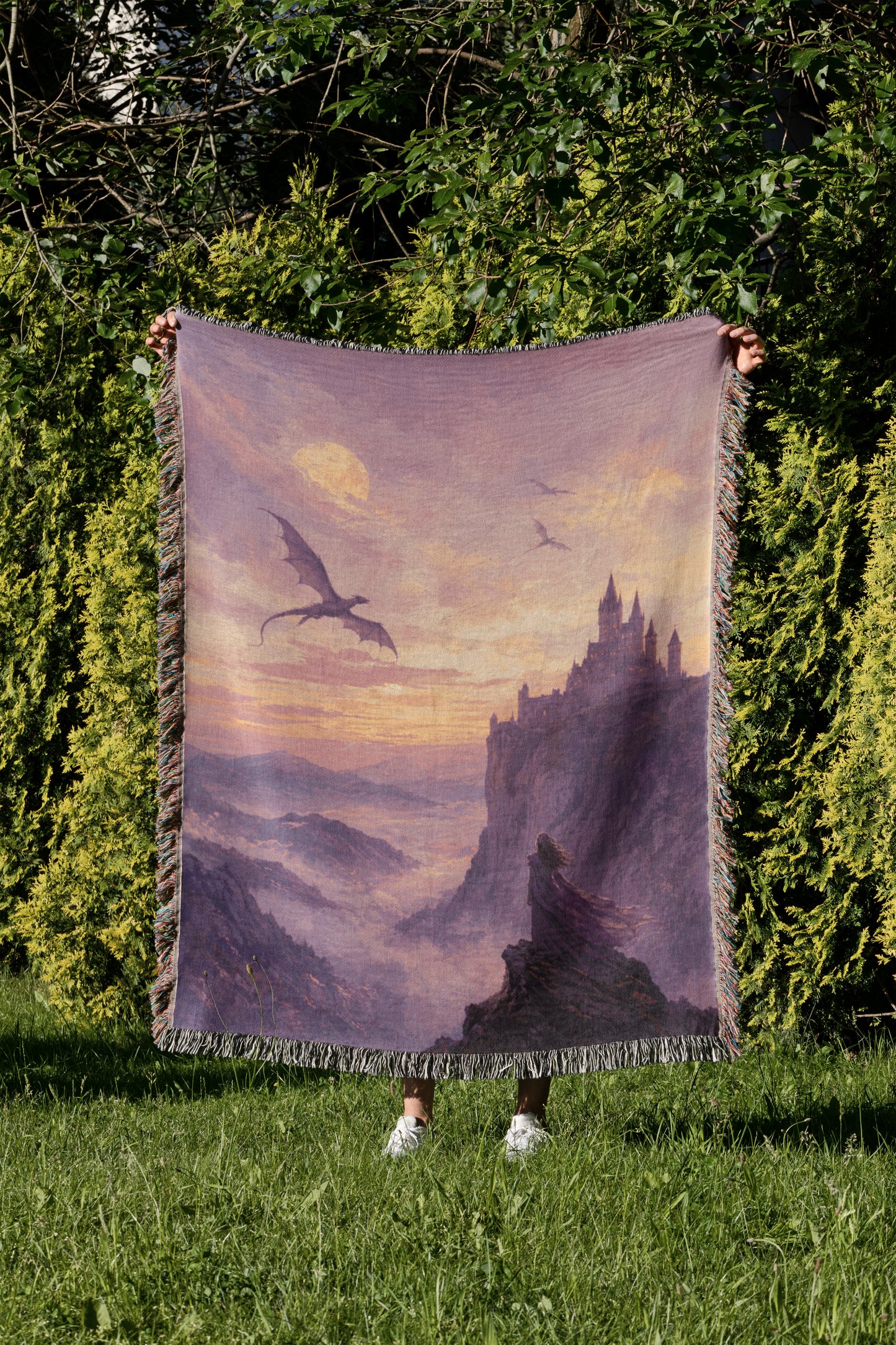 A person is standing in a grassy area, holding up a large blanket with a dragon and castle scene printed on it.