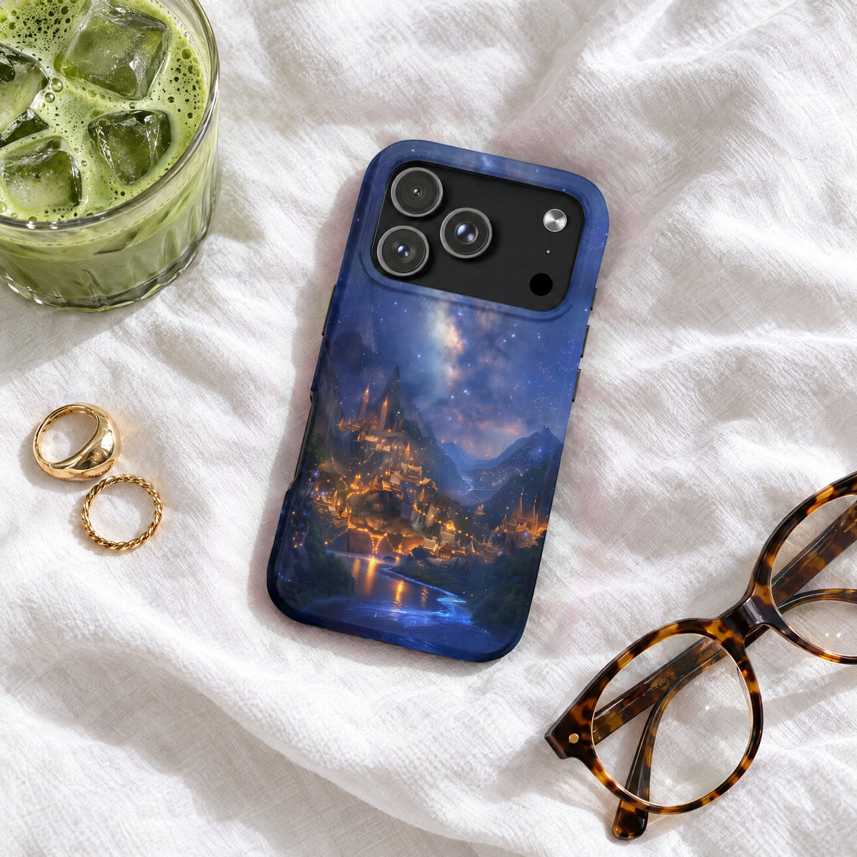 A blue phone case with a fantasy castle landscape design rests on a white sheet next to glasses, rings, and an iced drink.