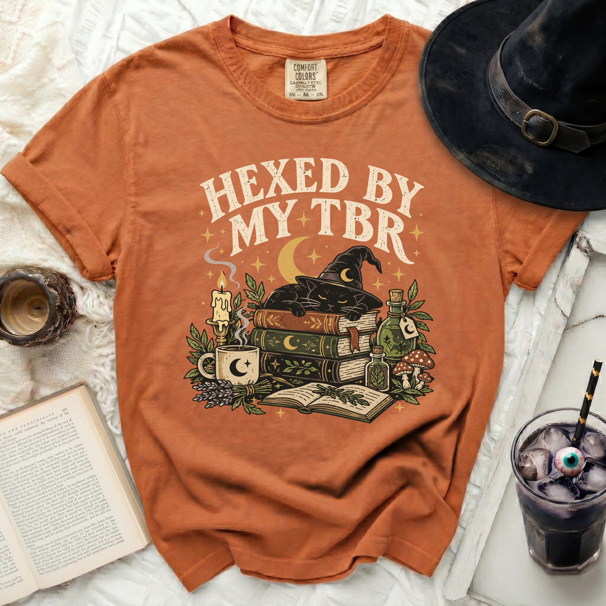 Orange t-shirt with a graphic of a black cat, books, candle, and potions, reading "Hexed by my TBR".