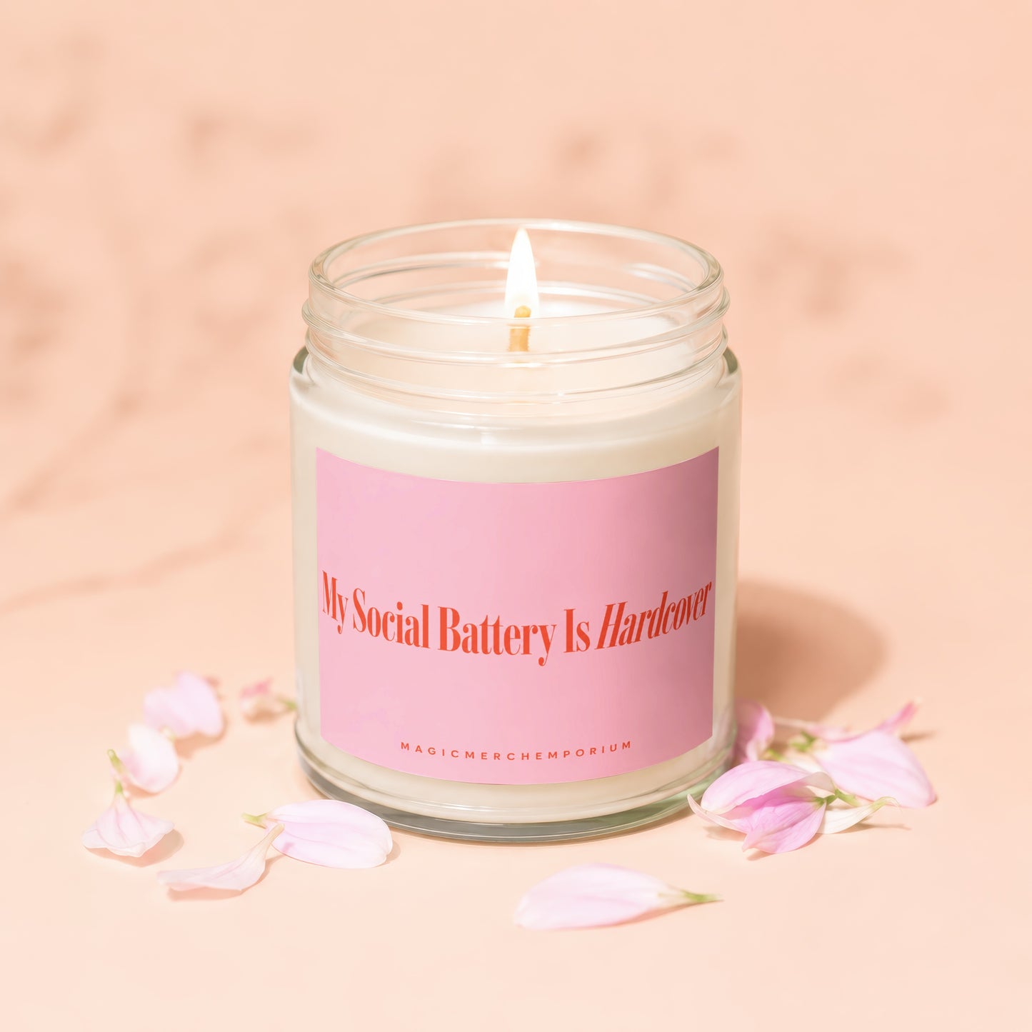 Glass jar candle with pink label reading "My Social Battery Is Hardcover" surrounded by petals.