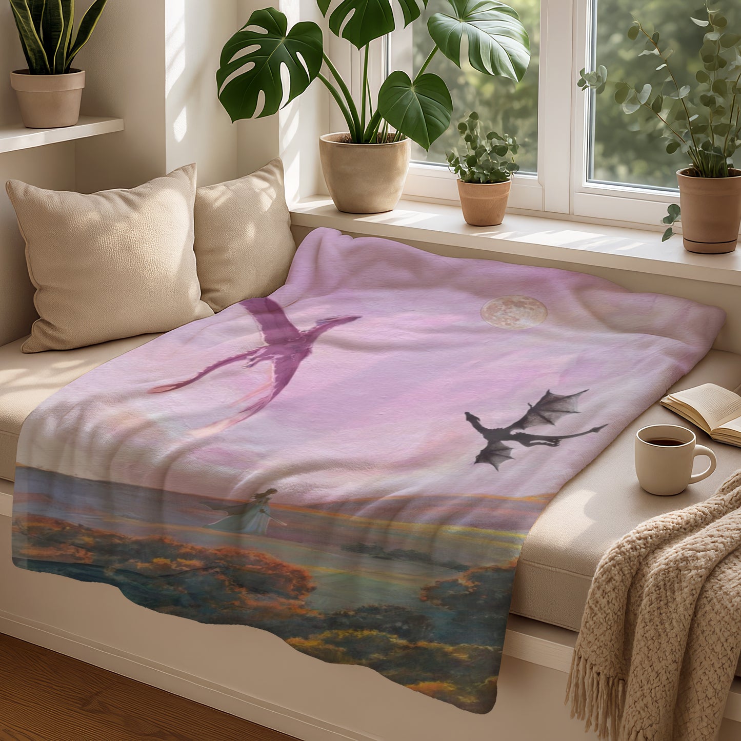 Pink blanket with dragon and moon artwork beside a book and mug.