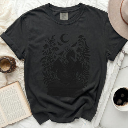 A black t-shirt features a graphic print of a dragon emerging from a teacup atop books, surrounded by foliage and a crescent moon.