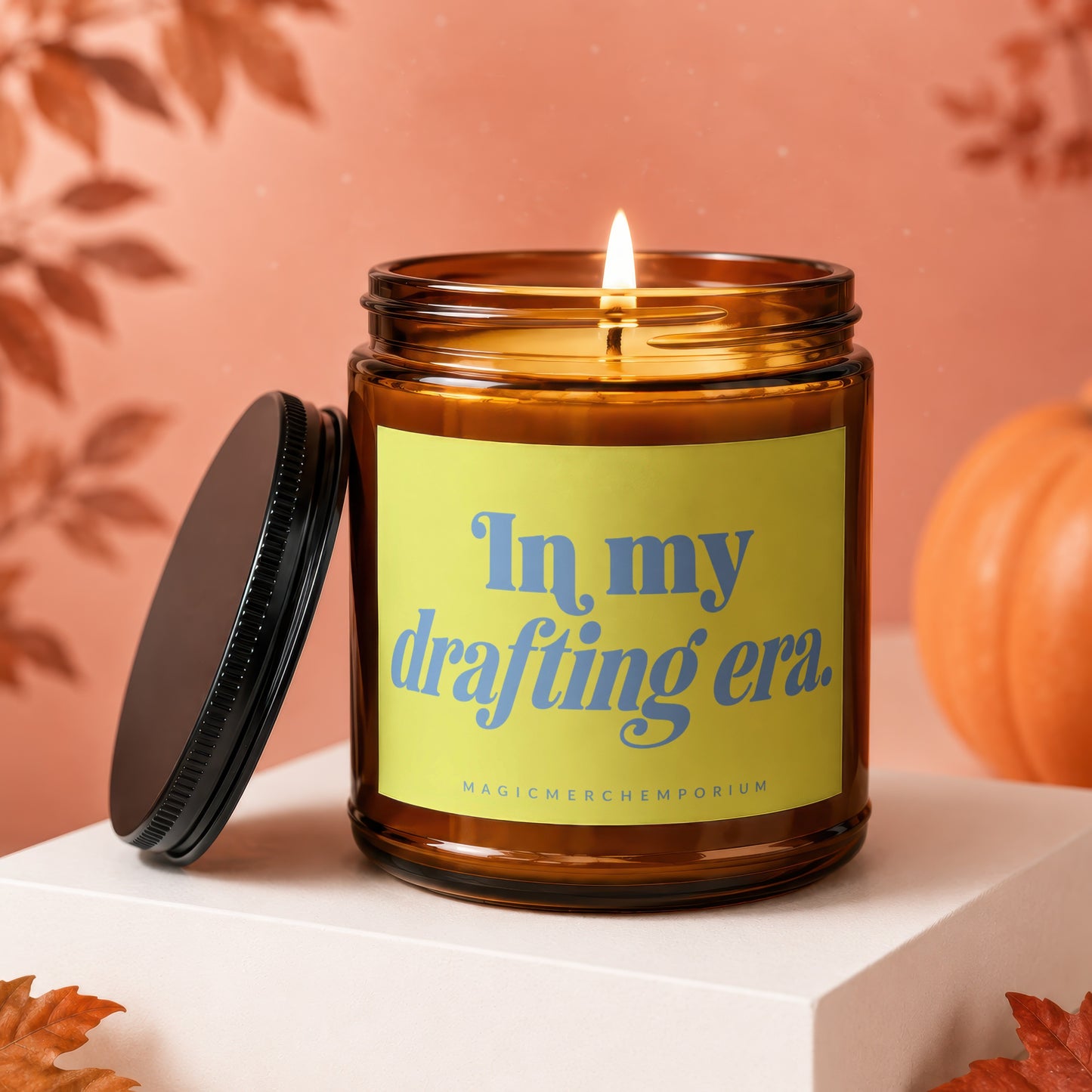 Amber glass candle with lit wick and yellow label reading "In my drafting era."
