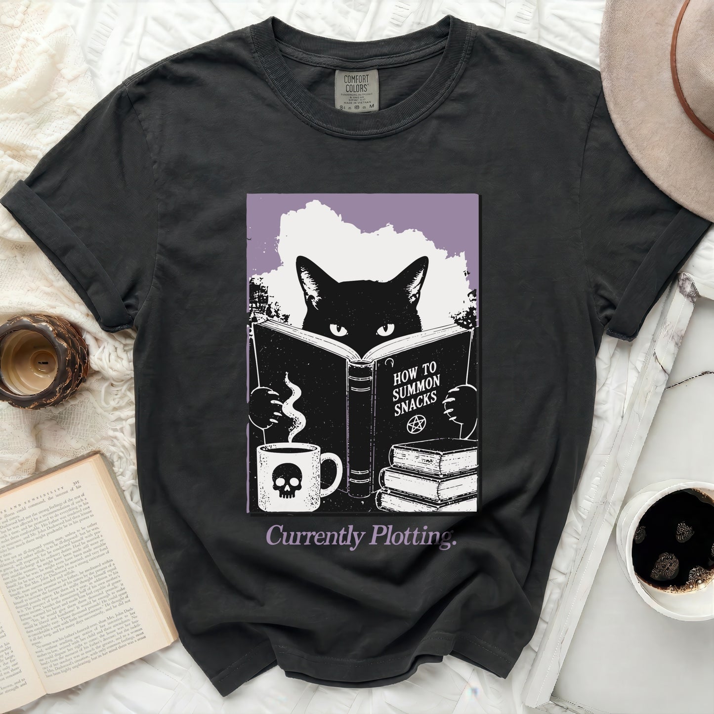 Black cat reading a book titled "How to Summon Snacks" with a skull mug.