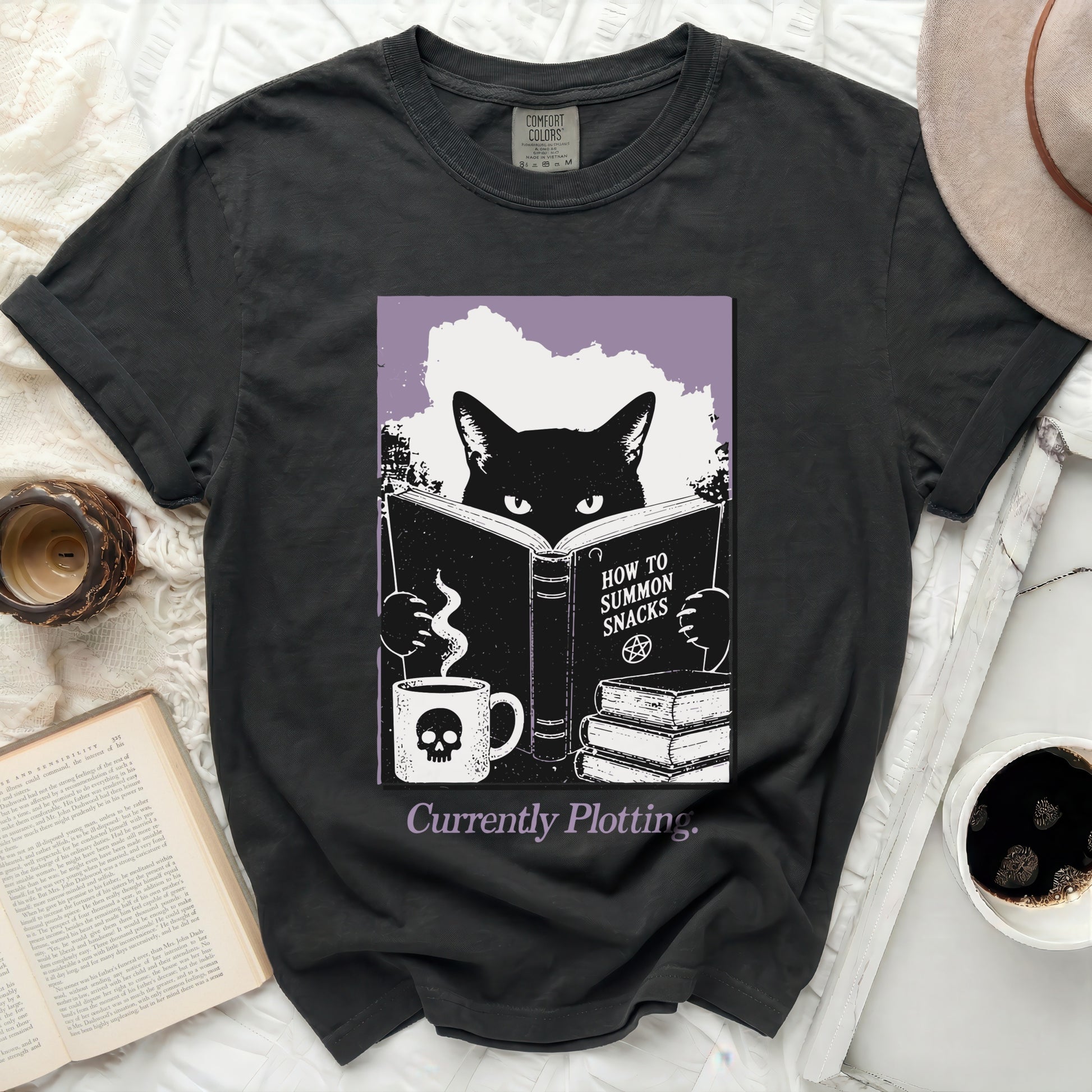 Black cat reading a book titled "How to Summon Snacks" with a skull mug.