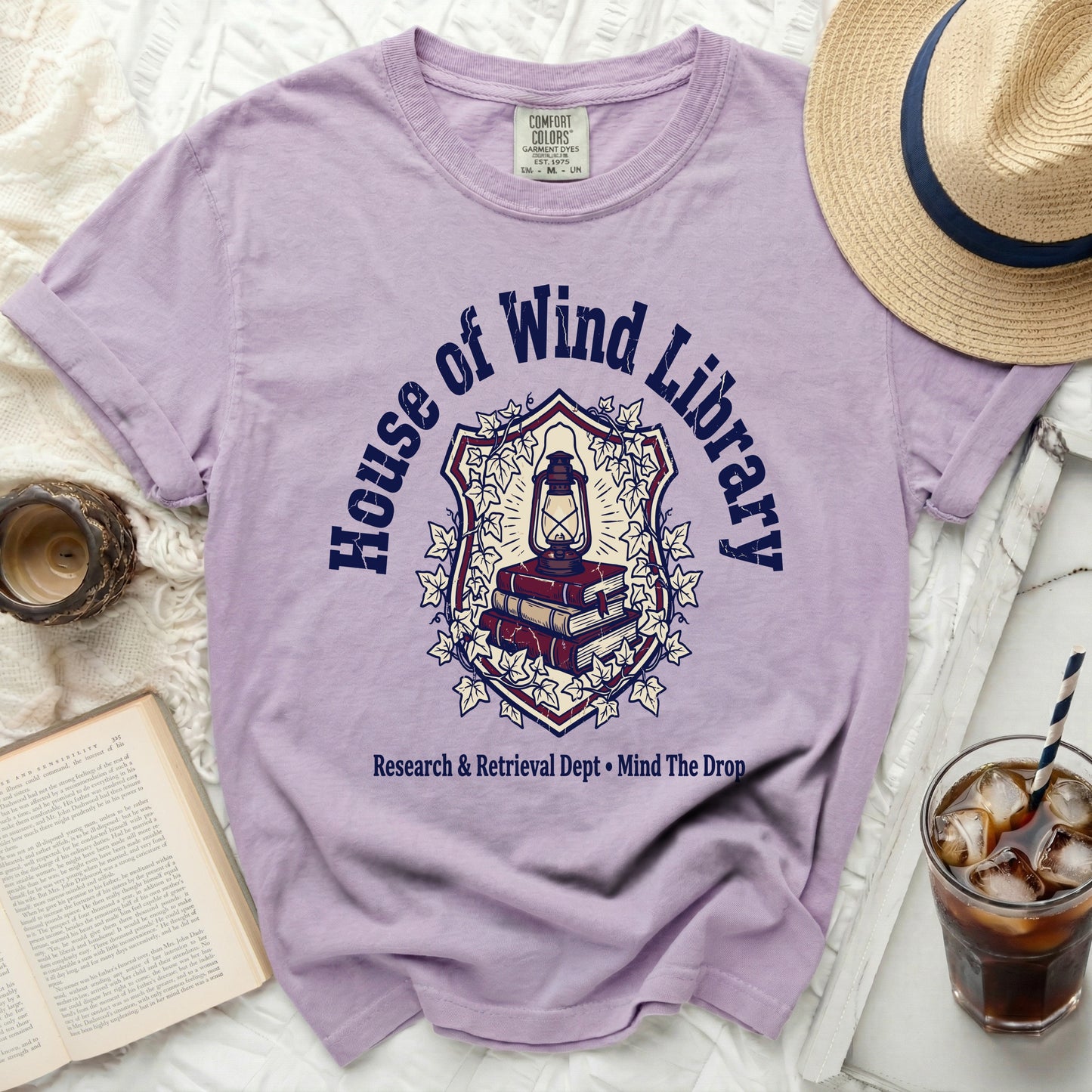 Purple t-shirt with "House of Wind Library" crest, lantern, and books graphic.