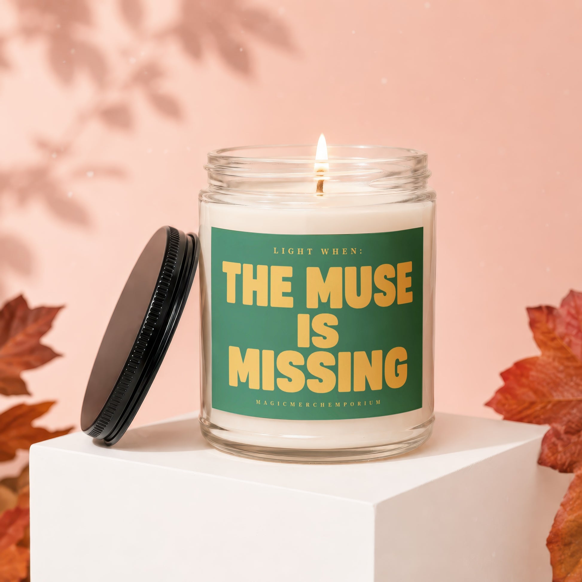 Glass candle with lit wick on white pedestal, "The Muse is Missing" text.
