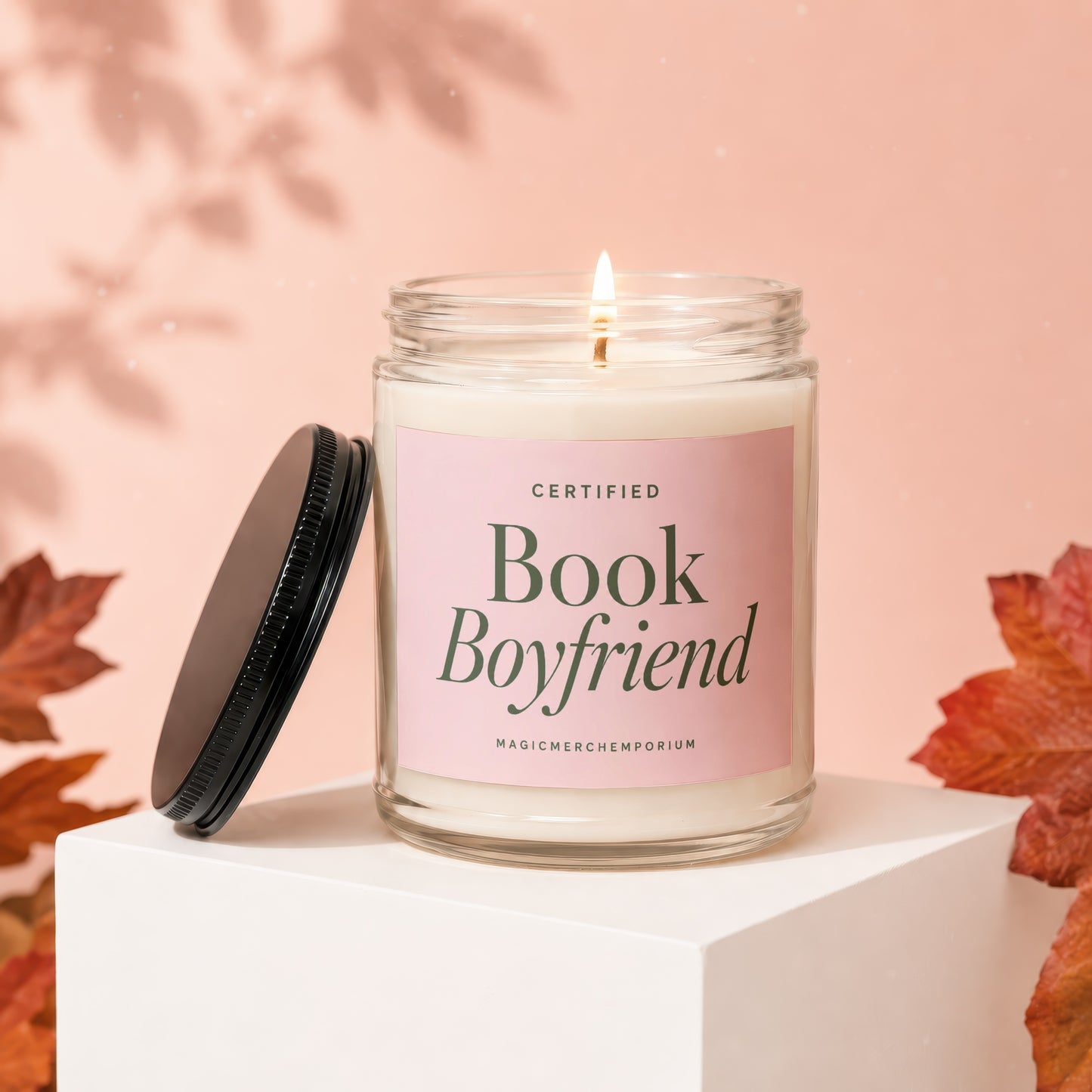 Candle with "Book Boyfriend" on pink label, lit, with lid off and autumn leaves.