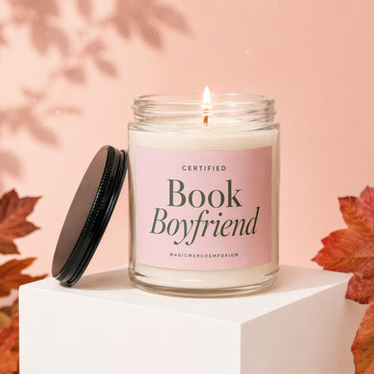 Candle with "Book Boyfriend" on pink label, lit, with lid off and autumn leaves.