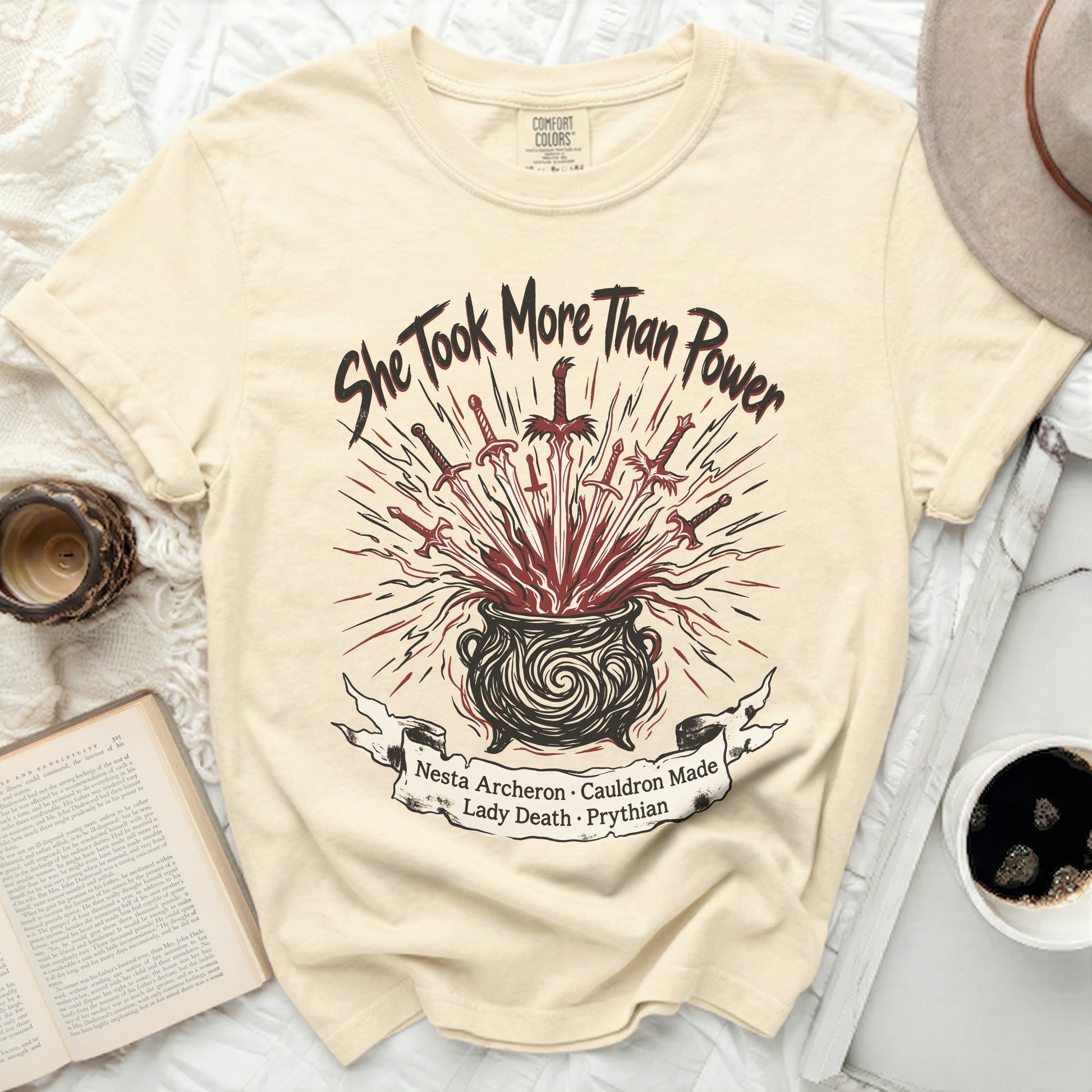 Cream t-shirt with "She Took More Than Power" and a cauldron with swords graphic.