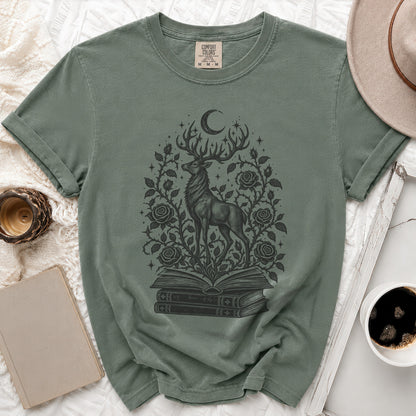 A sage green t-shirt displays a black graphic of a deer standing on stacked books surrounded by roses, vines, and a crescent moon.