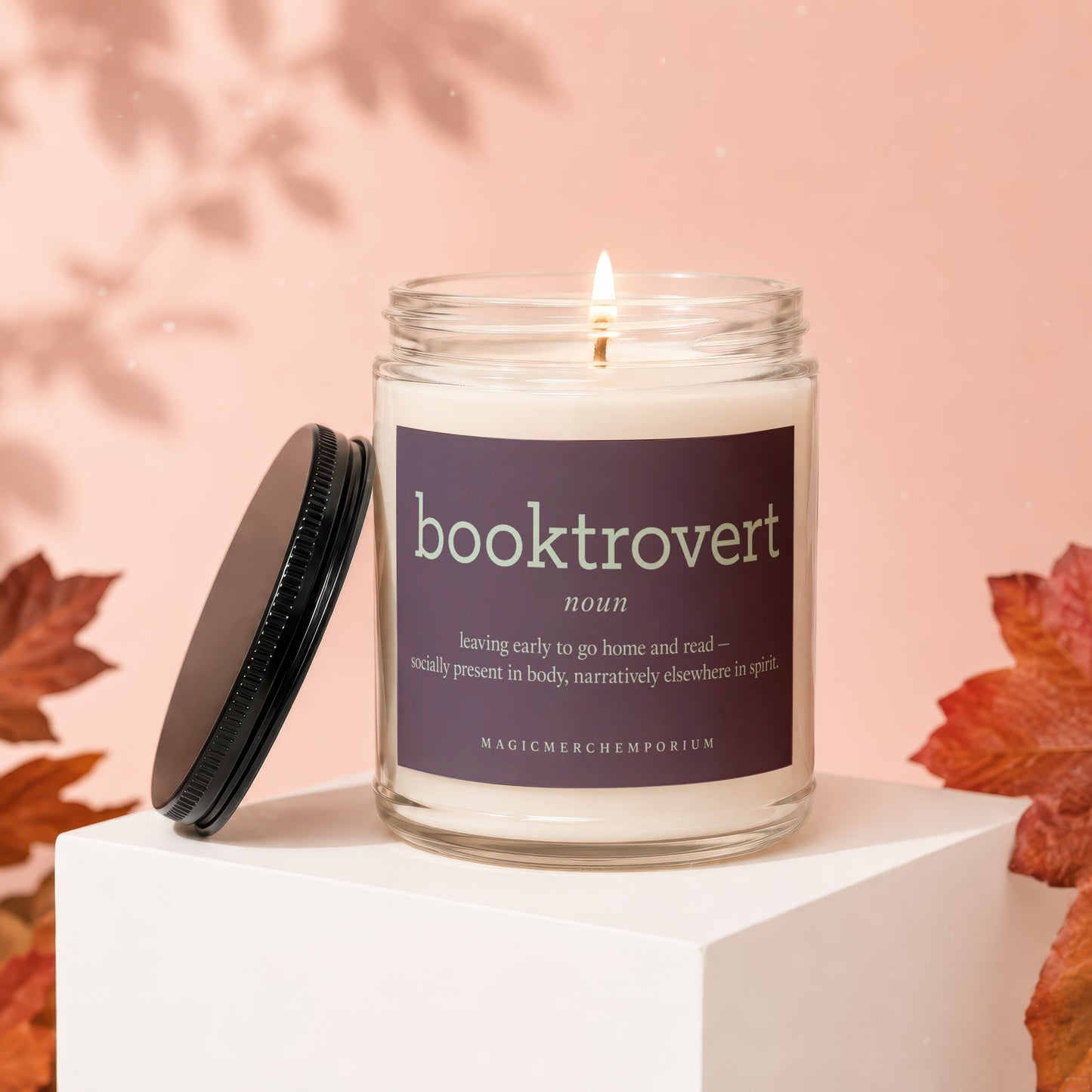 Lit candle with 'booktrovert noun' label, lid off, autumn leaves background.