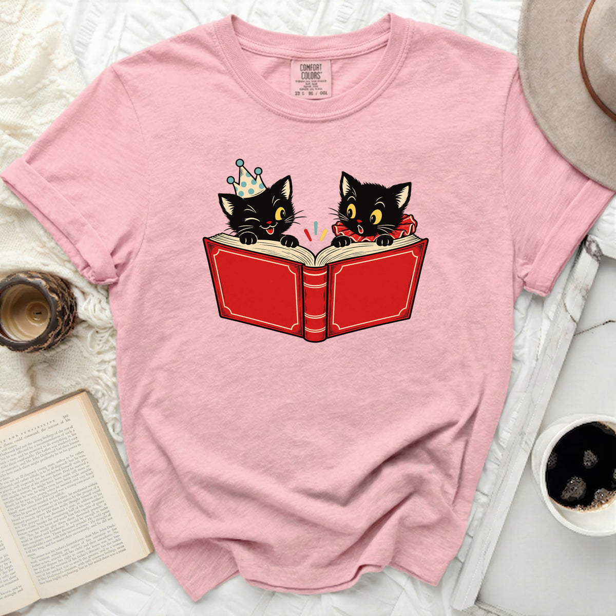 Pink t-shirt with illustration of two black cats reading a red book.