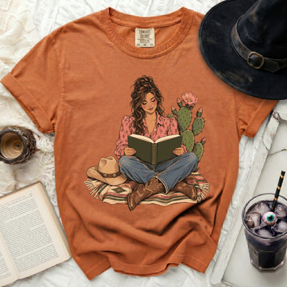 Orange Comfort Colors shirt featuring a graphic of a woman reading with cacti, a cowboy hat.