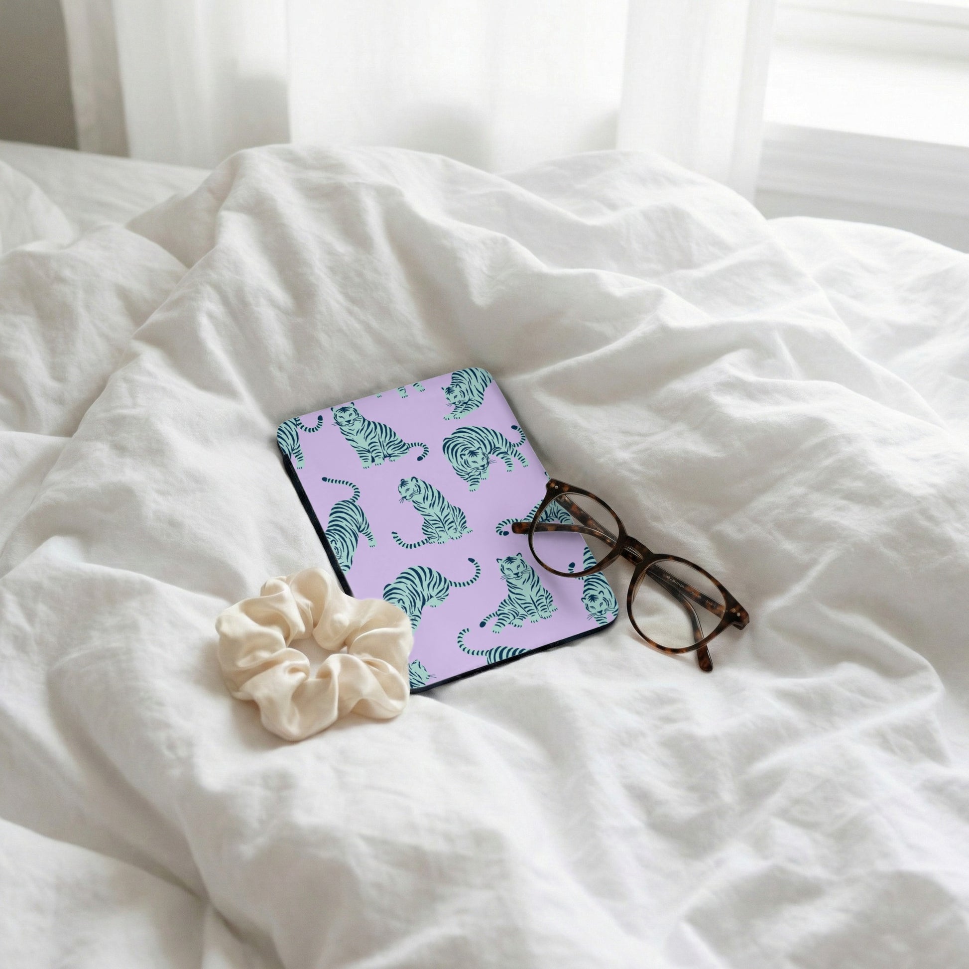 Purple case with teal tiger pattern, glasses, and scrunchie on white linens.