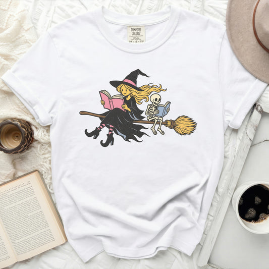 White t-shirt with illustration of witch and skeleton reading on a broomstick.