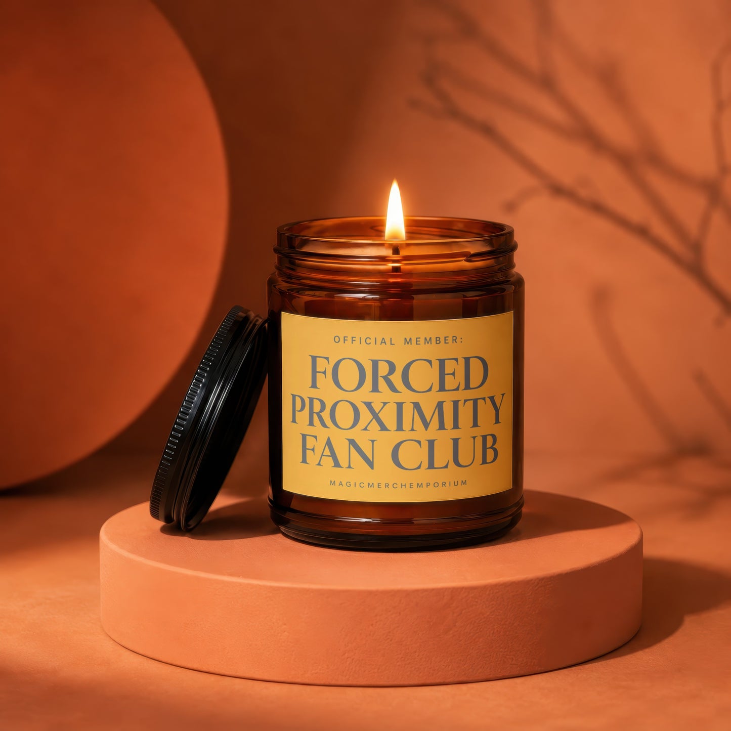 Amber candle with lit flame and black lid, reads "Forced Proximity Fan Club".