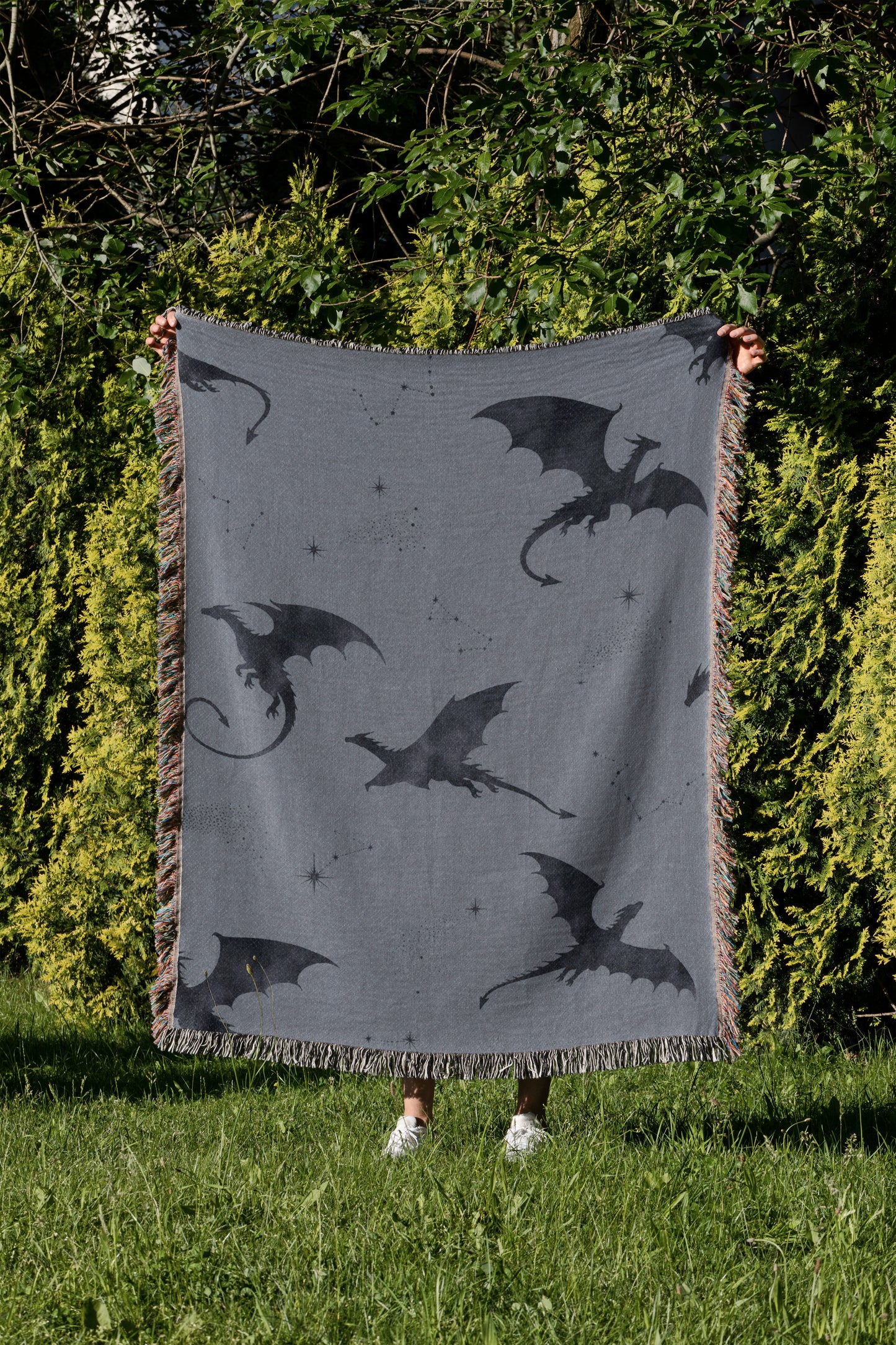 A person is holding up a gray blanket with black dragon silhouettes on it, standing in a grassy area with trees in the background.
