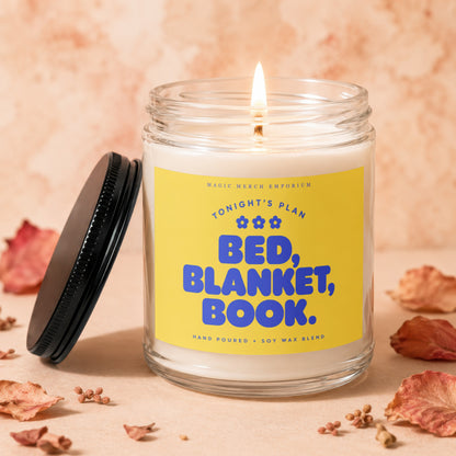 A lit candle in a glass jar with a yellow label reading "Bed, Blanket, Book."
