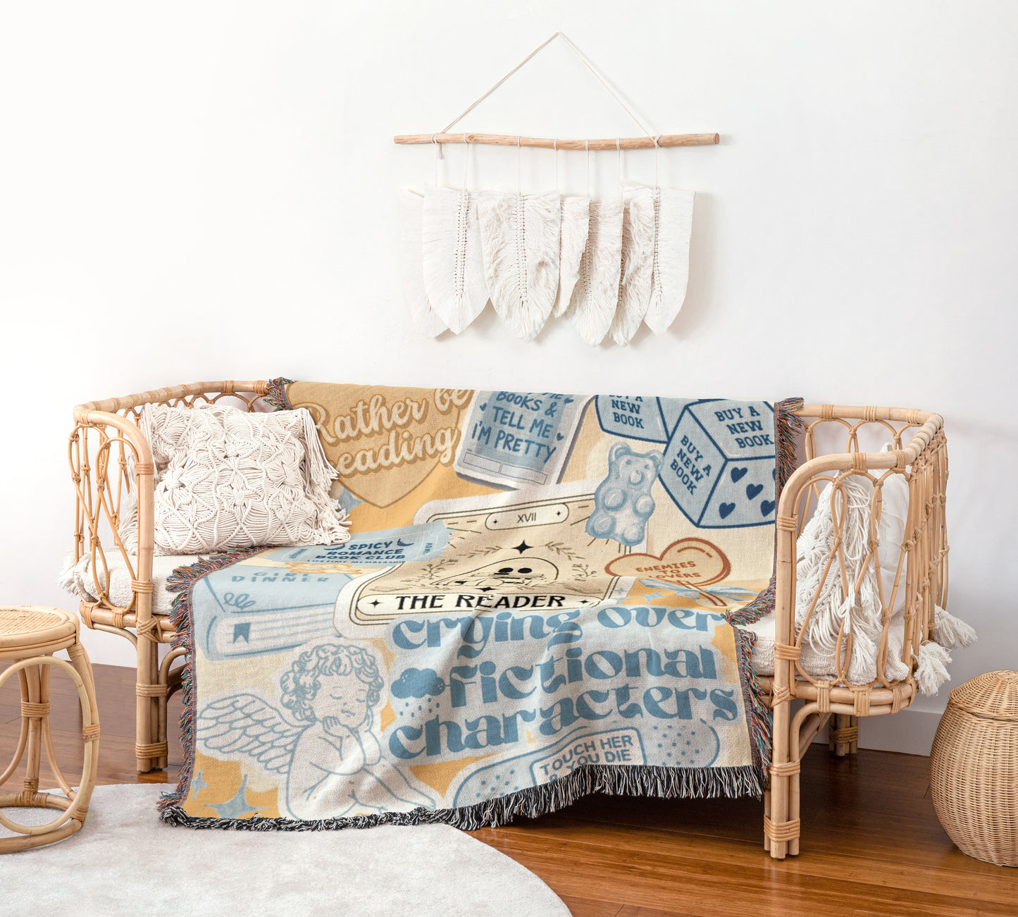 a cozy and inviting living space with a woven bench and a decorative wall hanging. The bench is adorned with a blanket that has a whimsical and playful design, including a character and the words "The Reader" and "Crying over fictional characters". The wall hanging is a collection of feathers, adding a touch of nature and rustic charm to the room. The overall atmosphere of the space is warm and inviting, with a harmonious blend of textiles and decorative elements.