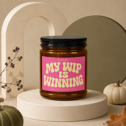 Amber jar candle with pink label reading: "STATUS: MY WIP IS WINNING"