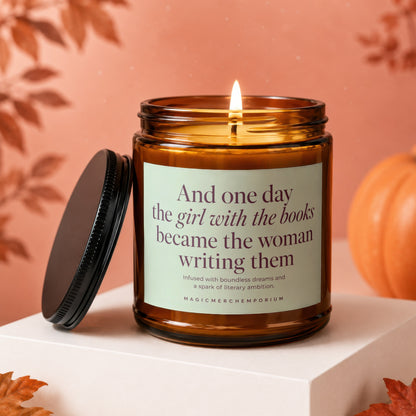 Amber glass candle with lit wick, featuring text "And one day the girl with the books became the woman writing them."